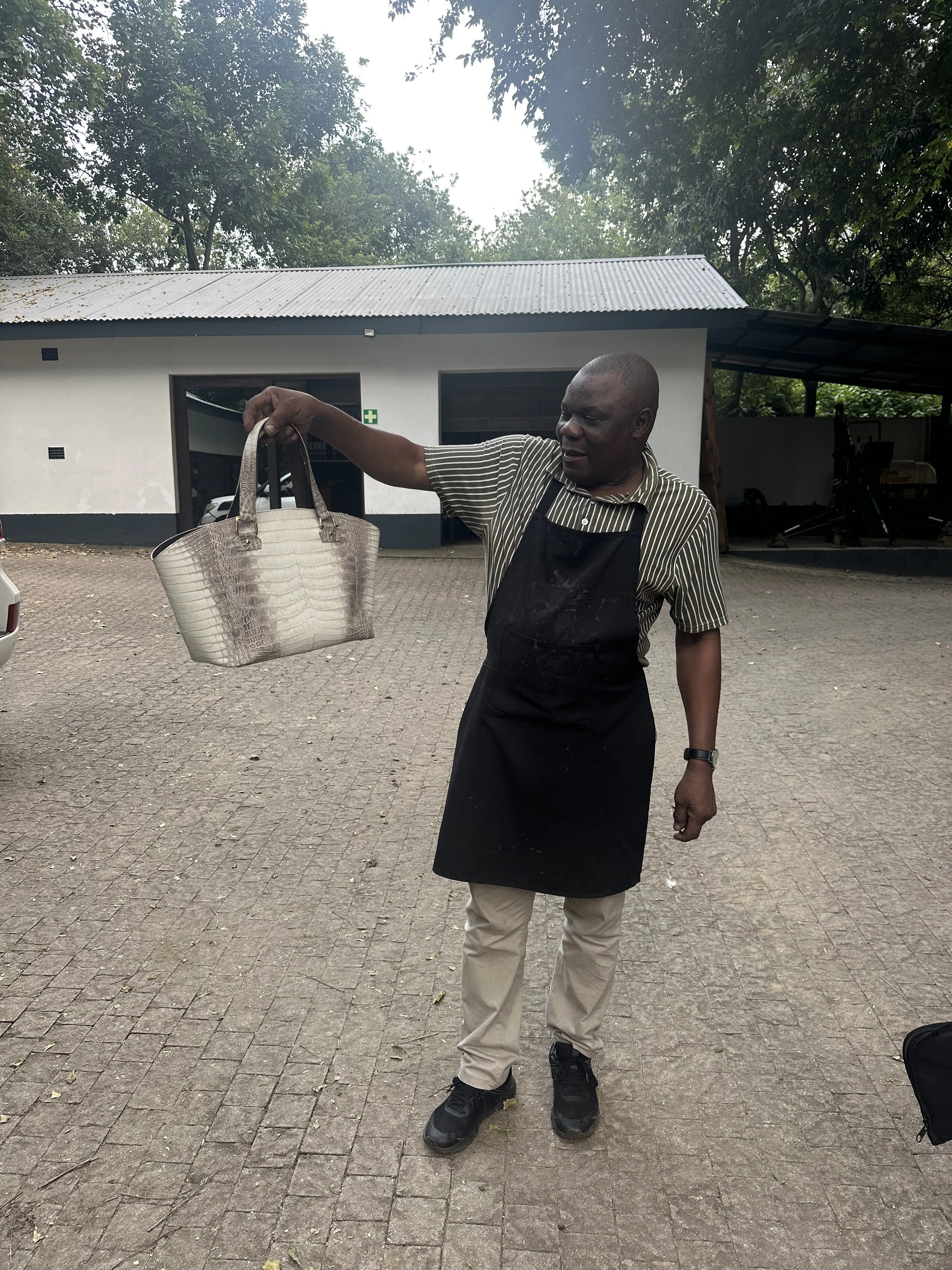 A maker in an apron holds up a finished crocodile leather handbag outdoors